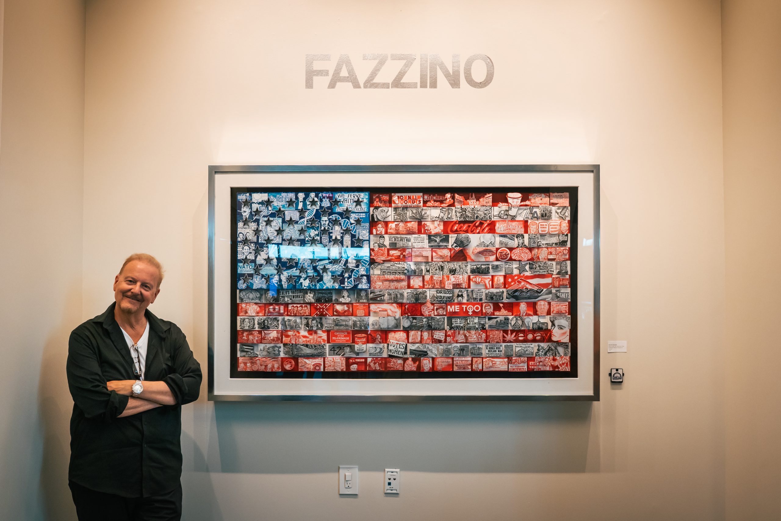 Fazzino stands in front of his American flag artwork, full of iconic historic moments in US history, smiling with his arms casually crossed