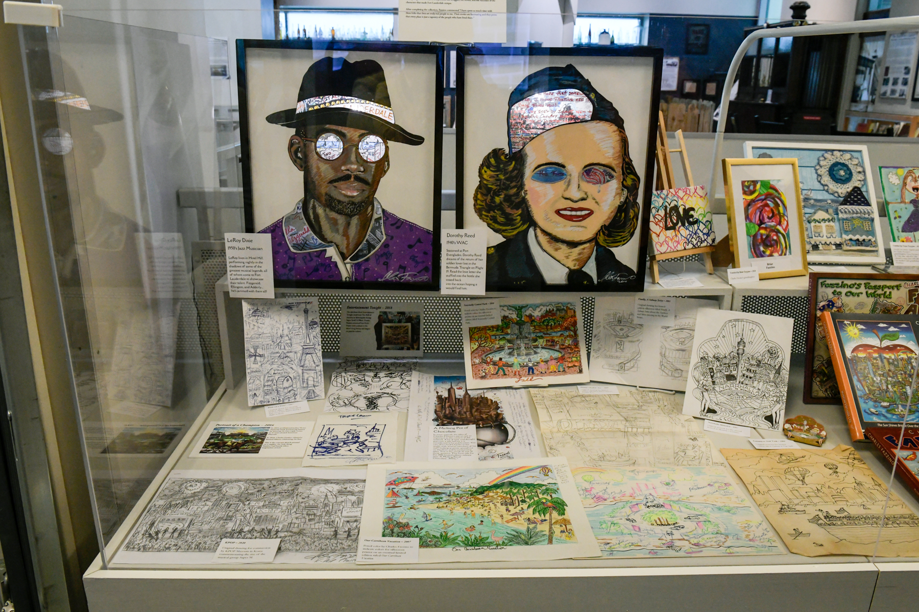 Two portraits, one of a man in a purple shirt and fedora, another of a women with a bob and military hat sit alongside other items from Fazzino's Faces of Ft. Lauderdale exhibit