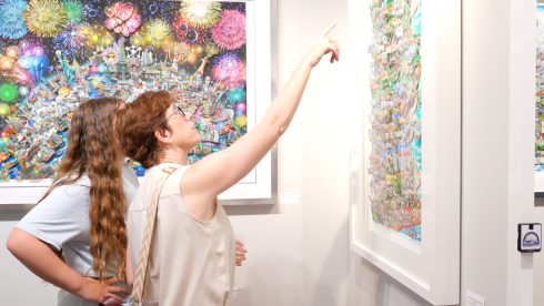 A person points at the top of a Fazzino piece hanging on the wall, her friend next to her